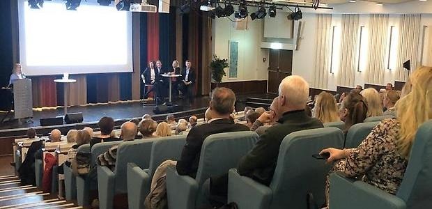 Många åhörare i en stor aula, en person presenterar från en pulpet och 4 personer sitter vid ett bord på en scen.