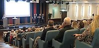 Många åhörare i en stor aula, en person presenterar från en pulpet och 4 personer sitter vid ett bord på en scen.
